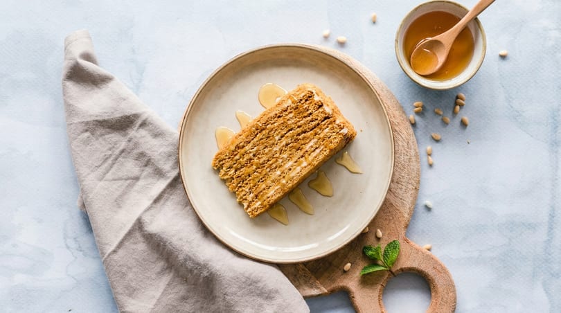Honey cake slice with honey drizzle on a plate, with a bowl of honey and pine nuts.