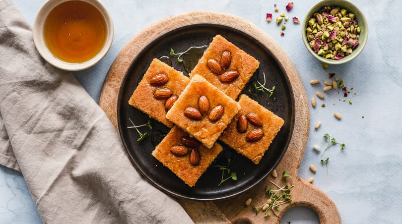 Overhead view of five square semolina cakes with almonds and honey, alongside honey, pistachios, and pine nuts.
