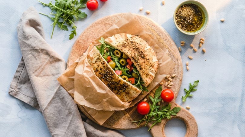 Pita bread sandwich filled with arugula, olives, and tomatoes, served with a side of olive oil.